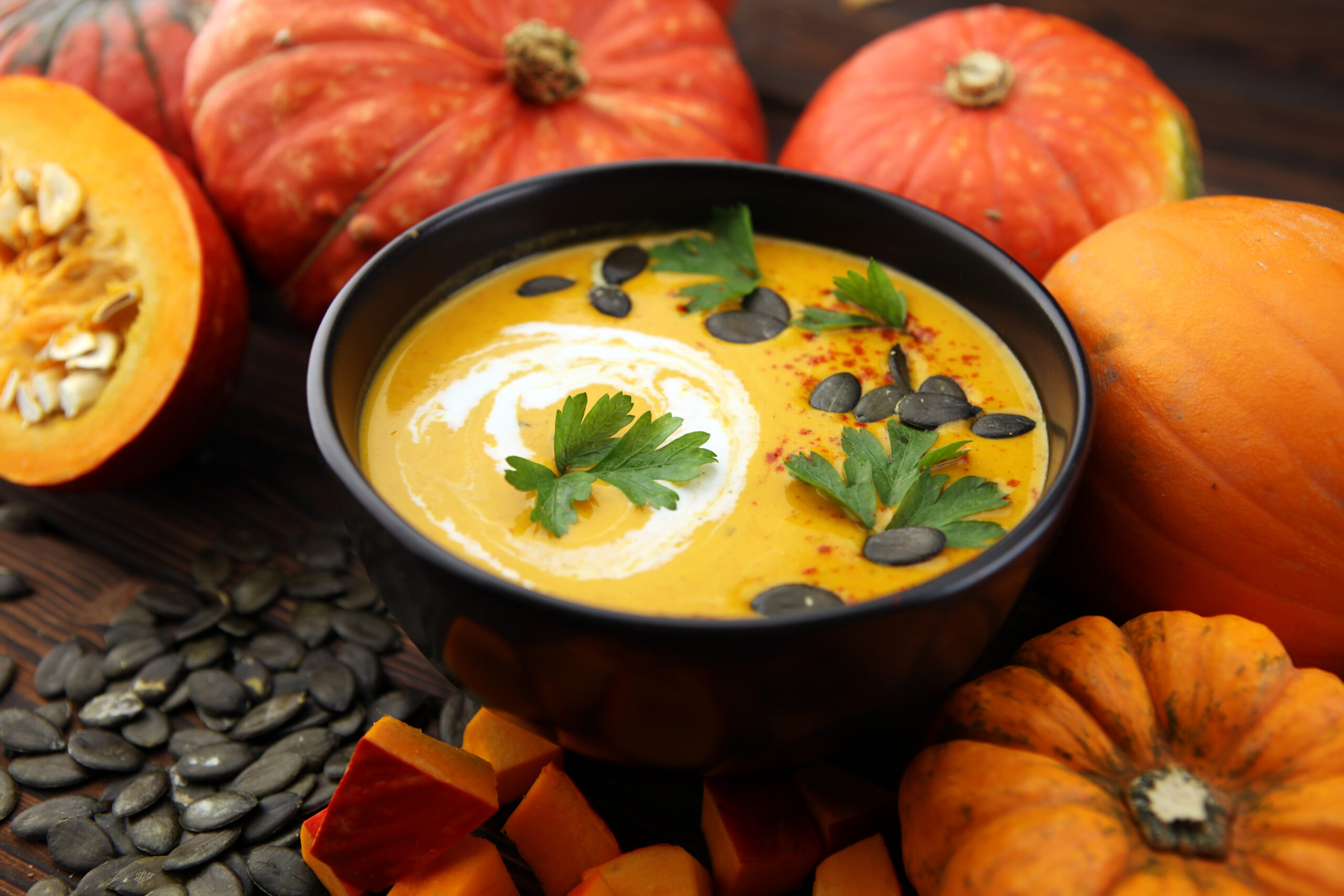 Eine Schüssel mit cremiger Kürbissuppe, garniert mit Kürbiskernen, Petersilie und einem Sahnetupfer - perfekt für das Immunsystem stärken - umgeben von ganzen Kürbissen, Kürbisstücken und Kürbiskernen.