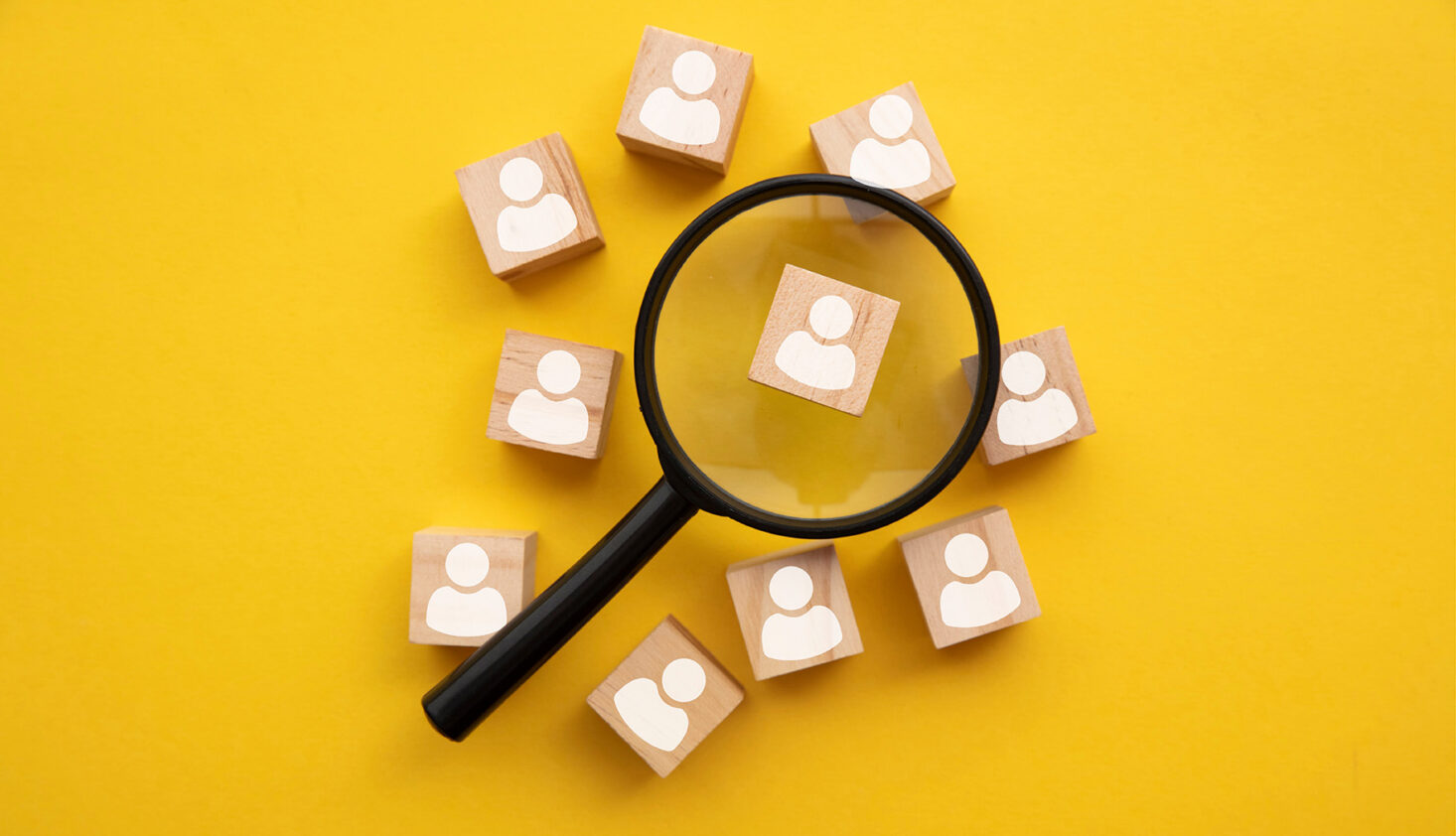 Eine Lupe, die auf einen Holzblock mit einem Benutzersymbol gerichtet ist, umgeben von ähnlichen Blöcken, alle auf gelbem Hintergrund, symbolisiert die Bedeutung von Lebenspflege und individueller Definition.