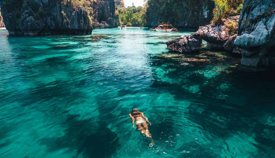 Eine Person genießt ihren Urlaub beim Schwimmen im klaren türkisfarbenen Wasser, umgeben von felsigen Klippen und üppigem Grün, mit kleinen Booten, die in der Ferne unter einem hellen, sonnigen Himmel schwimmen - perfekt, um gesund zu reisen und Krankheiten vorzubeugen.
